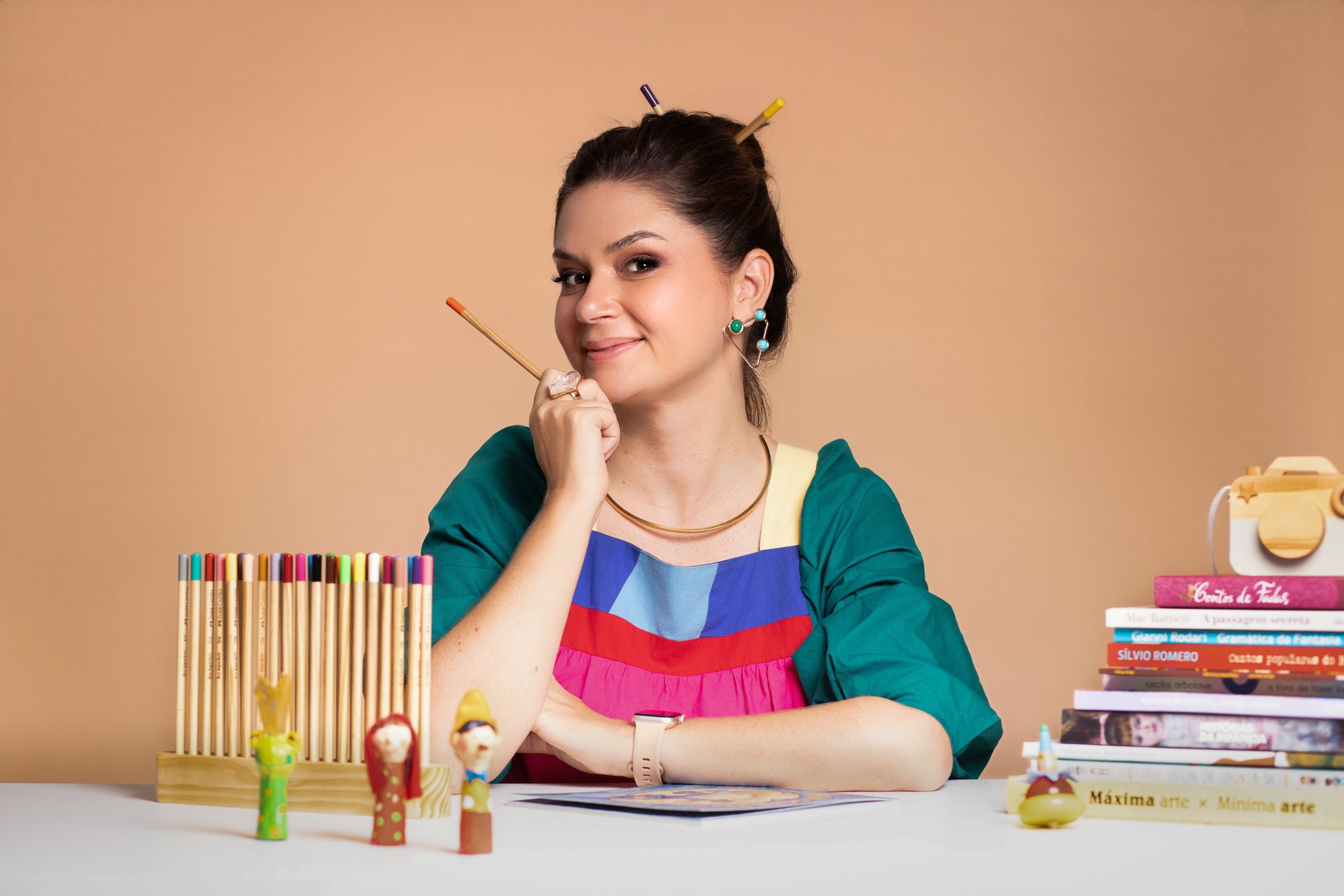 Aline Pinto sentada à mesa, sorrindo, com lápis de cor no cabelo, cercada de livros e materiais de arte